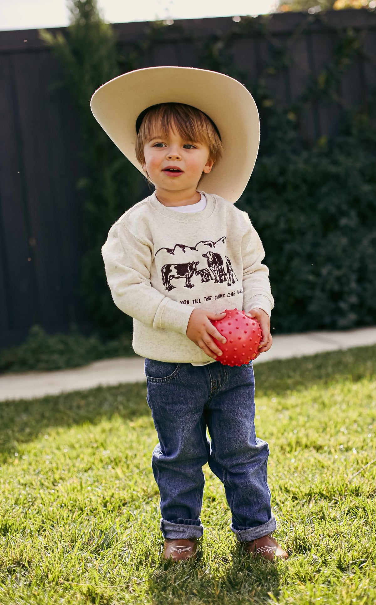 Picture of a toddler boy standing with a sand color crewneck sweatshirt on with a scene of 3 cows and a baby cow embroidered on the front of the sweatshirt with the words Love You Till The Cows Come Home Written underneath.