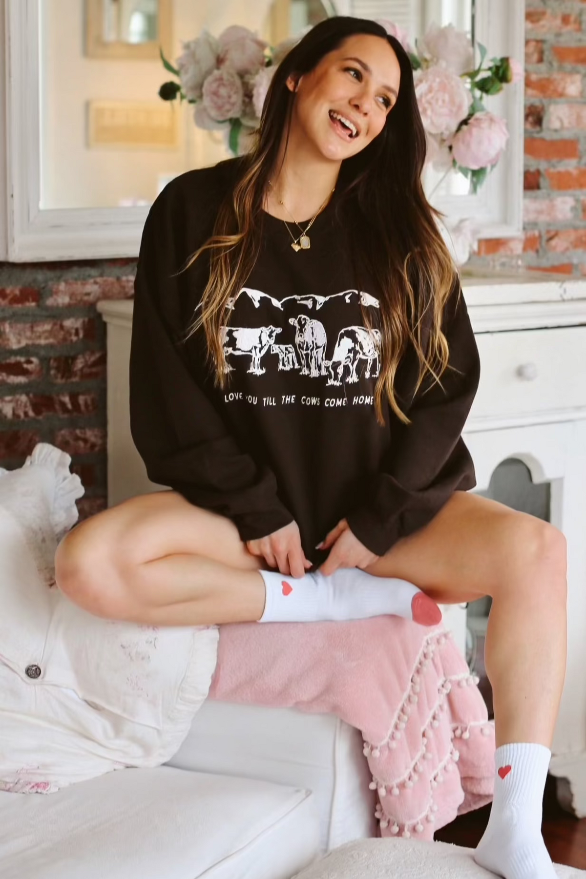 Picture of brunette model sitting on a couch with a brown crewneck sweatshirt on that has a scene of 3 cows and a baby cow in white embroidery with the words Love You Till The Cows Come Home underneath the scene.