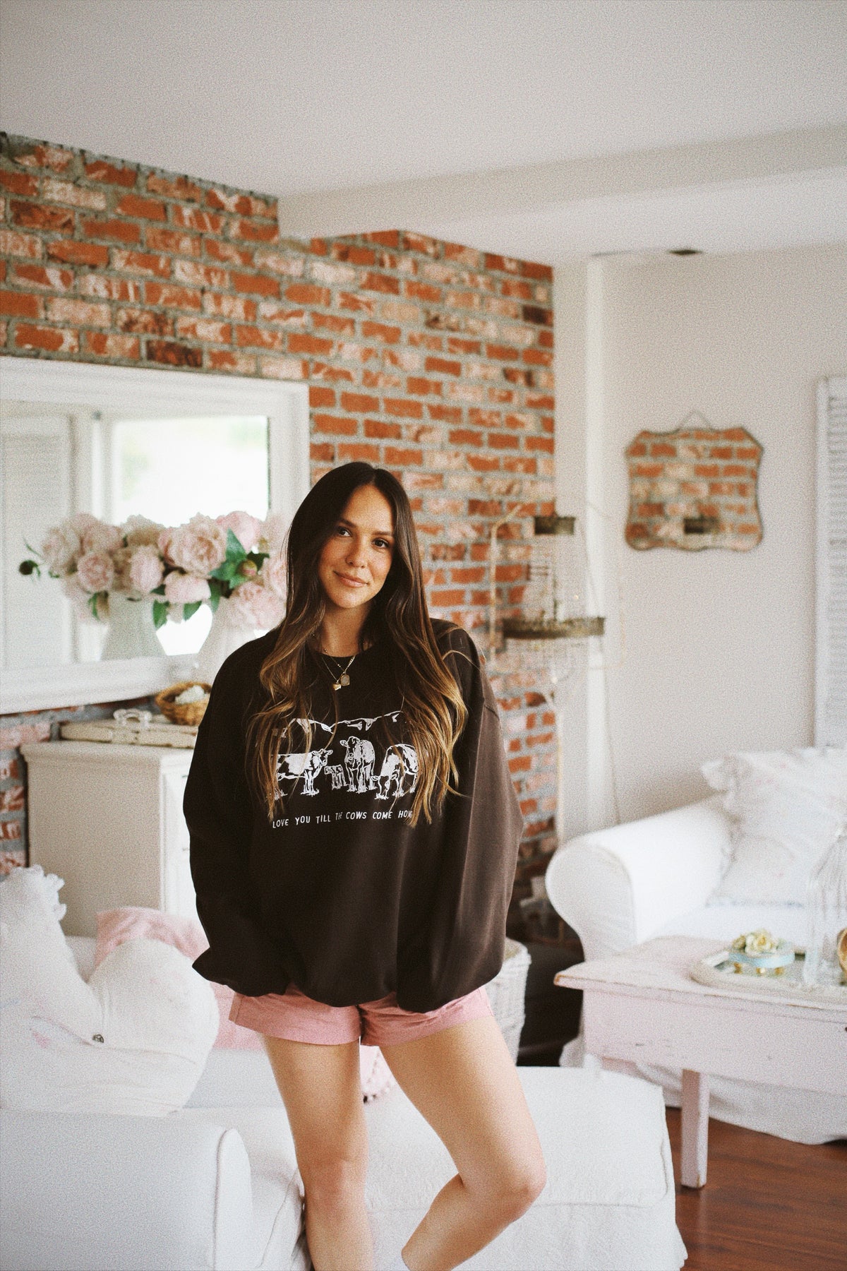 Picture of brunette model standing next to  a couch with a brown crewneck sweatshirt on that has a scene of 3 cows and a baby cow in white embroidery with the words Love You Till The Cows Come Home underneath the scene.