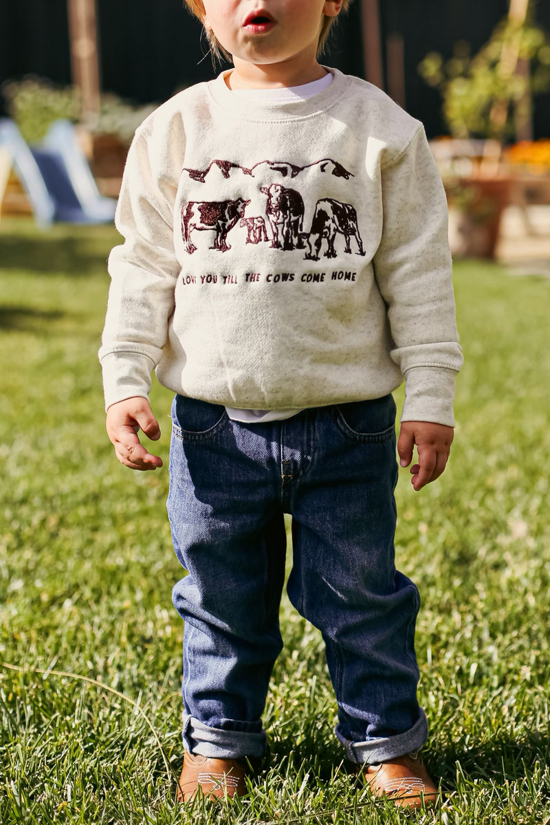 Picture of a toddler boy standing with a sand color crewneck sweatshirt on with a scene of 3 cows and a baby cow embroidered on the front of the sweatshirt with the words Love You Till The Cows Come Home Written underneath.