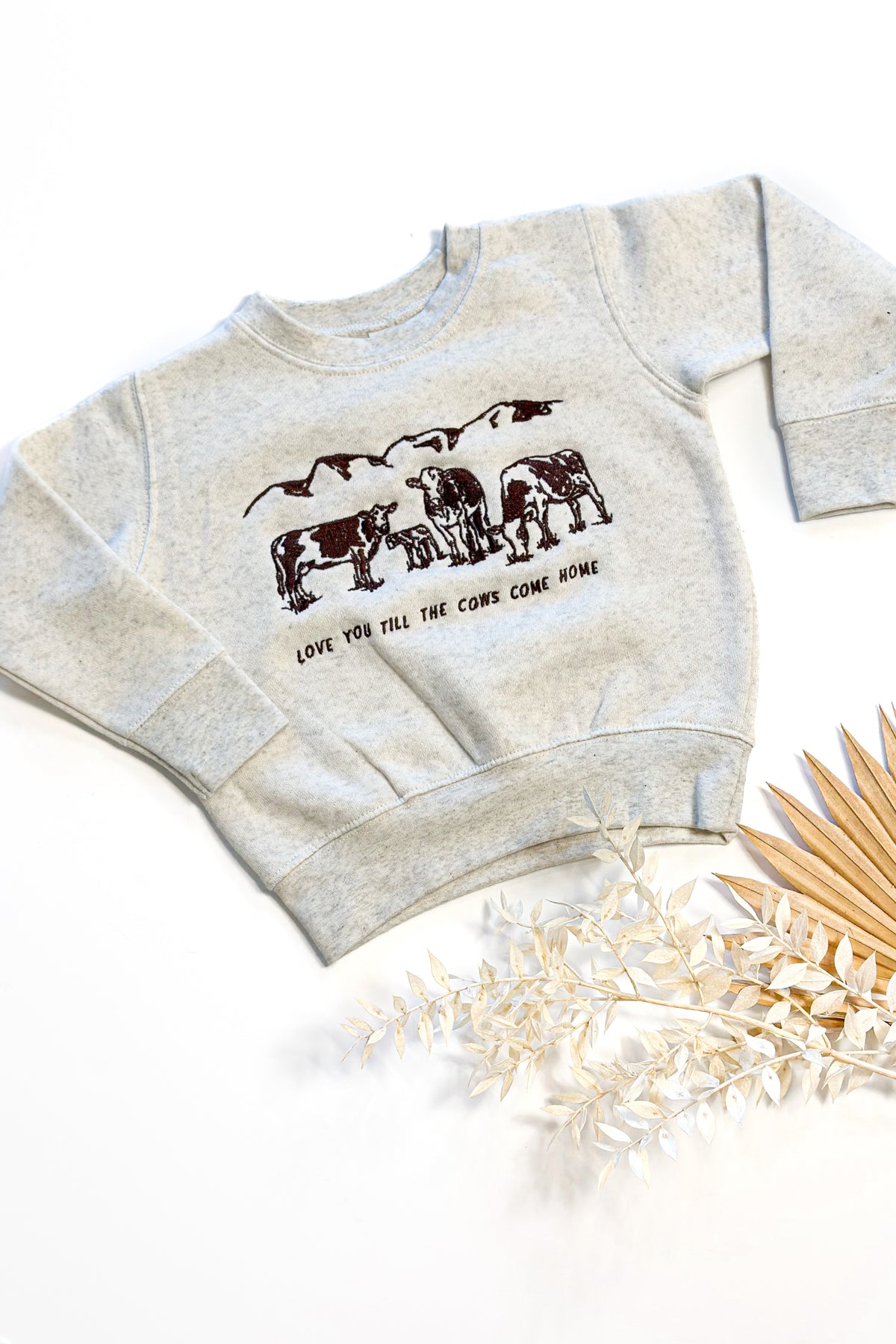 Picture of a sand color crewneck sweatshirt on with a scene of 3 cows and a baby cow embroidered on the front of the sweatshirt with the words Love You Till The Cows Come Home Written underneath.