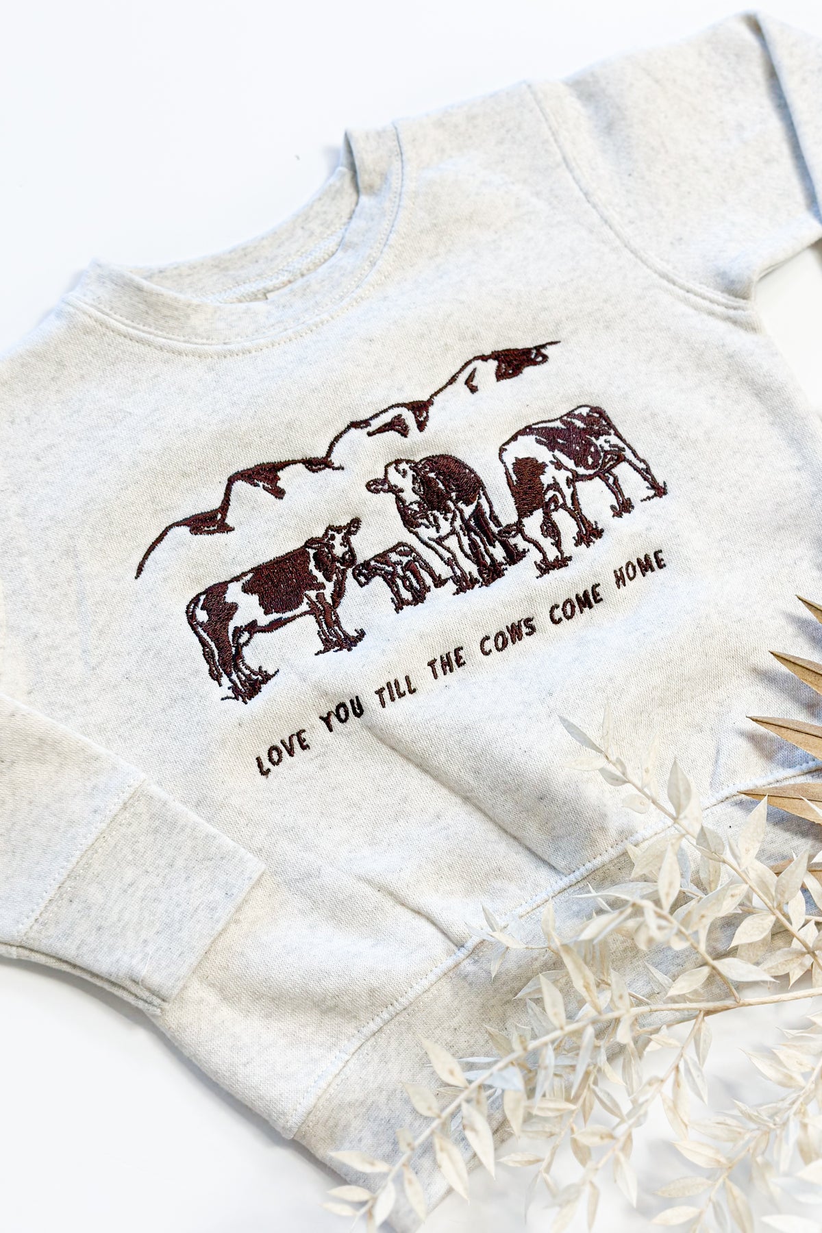 Picture of a sand color crewneck sweatshirt with a scene of 3 cows and a baby cow embroidered on the front of the sweatshirt with the words Love You Till The Cows Come Home Written underneath.